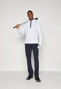 Polo blanco de manga larga con media cremallera, combinado con pantalones navy y zapatos negros. Sostiene un palo de golf y lleva un guante de golf blanco en la mano izquierda.