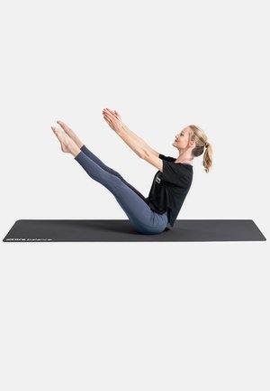 Black exercise mat with a woman performing a seated V-up, wearing a black t-shirt and blue leggings. Focus on core strength and balance.