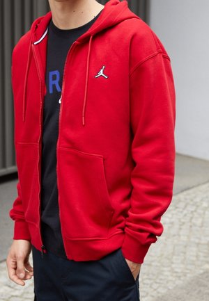 Person iført en rød hættetrøje med lynlås og et hvidt og sort logo på brystet, en sort t-shirt og mørke bukser med den ene hånd i lommen.