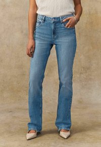 Jeans de mezclilla de color azul claro, acampanados y de cintura alta, con bolsillos frontales y un ligero desgaste. Combinados con tacones de punta beige.