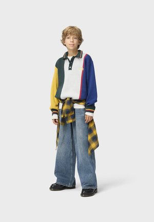 Kind met warrig haar dat een color-block lange mouwen polo draagt, wijde spijkerbroek, zwarte schoenen en een geel geruit overhemd om de taille gestrikt.