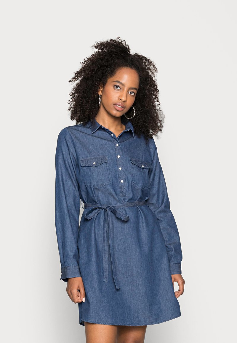 Vestido camiseiro em denim com mangas longas, frente abotoada, dois bolsos no peito, um cinto na cintura e uma bainha arredondada. Usado numa cor azul escura sólida.