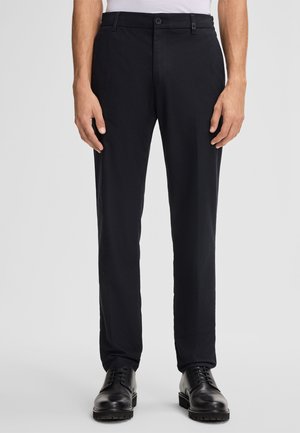 Schwarze Anzughose, schlanke Passform, aus gewebtem Stoff. Mit flacher Vorderseite, Gürtelschlaufen und zwei Seitentaschen. Kombiniert mit schwarzen Schuhen.