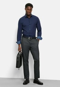 Marineblaue Langarmbluse mit gemustertem blauen Bund, graue strukturierte Hose, schwarzer Gürtel und Lederaktentasche, kombiniert mit schwarzen Schuhen.