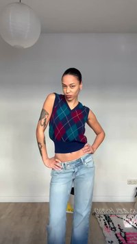 Γυναίκα με τατουάζ, φορώντας αμάνικο πλεκτό γιλέκο με ρόμβους και ανοιχτόχρωμο τζιν, στέκεται με τα χέρια στη μέση σε ένα μινιμαλιστικό δωμάτιο.