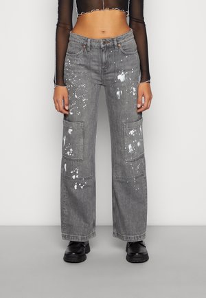 Vrouw draagt een high-waist grijze wijd uitgesneden jeans met witte verfspatten, grote kniezakken, zwarte chunky schoenen en een doorschijnende cropped top met lange mouwen.
