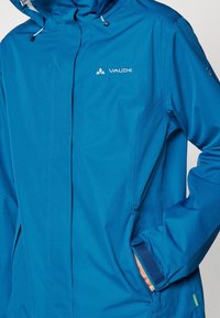 Veste imperméable bleue avec capuche, en tissu texturé, dotée d'une fermeture éclair avant, de cordons de serrage réglables et de poignets à scratch.