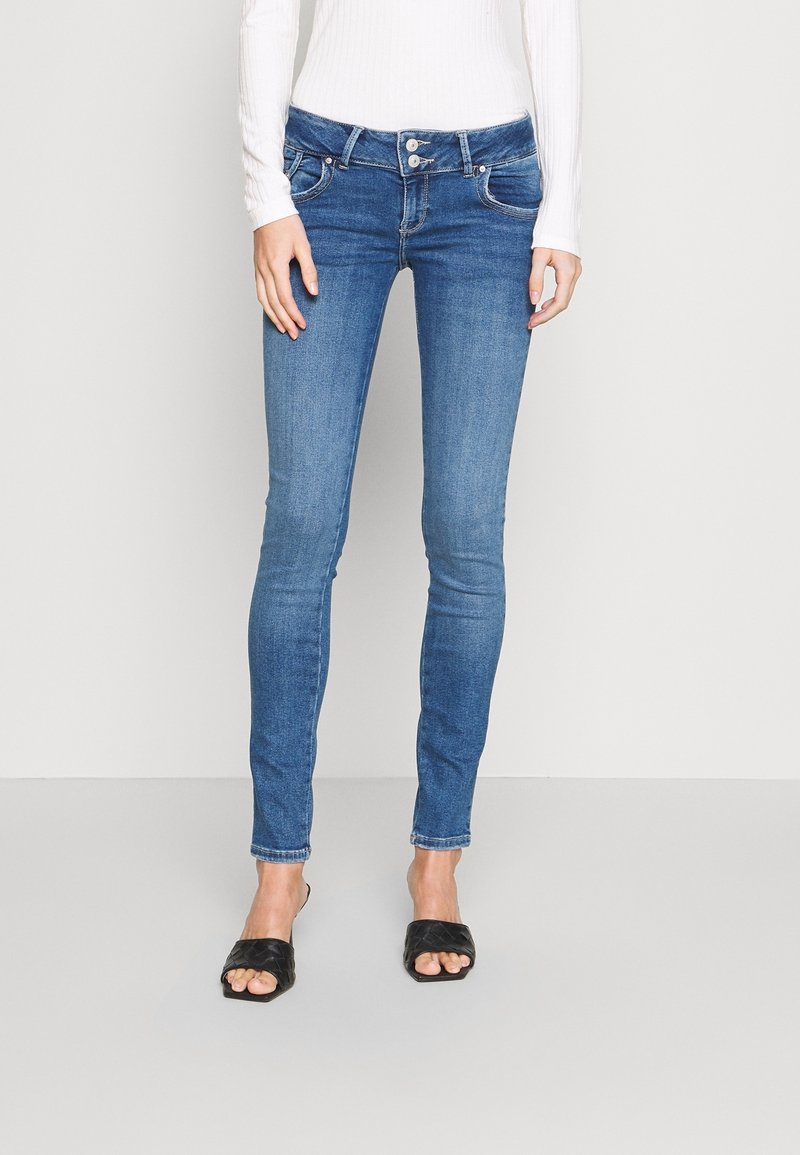 Femme portant un jean skinny bleu moyen avec une taille à double bouton et des sandales à talons noires à bout ouvert, se tenant devant un fond clair uni.