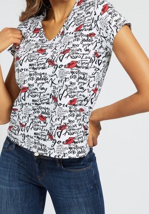 Frau trägt ein weißes V-Neck-Shirt mit schwarzem Schriftzug und roten Lippenabdrücken, kombiniert mit blauen Jeans, eine Hand in der Hüfte und die andere Hand am Kragen.