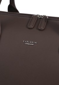 Smeđa kožna torba s glatkom teksturom, koja ima dvostruke rastezače i metalnu oznaku s natpisom "Carlheim Copenhagen."