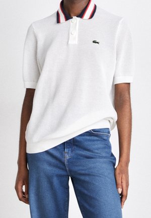 Persoon draagt een wit gestructureerd polohemd met korte mouwen en rood-wit-blauwe strepen op de kraag, en een blauwe spijkerbroek, staande tegen een effen achtergrond.