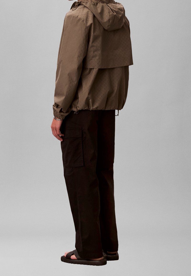 Personne portant une veste coupe-vent à capuche marron, un pantalon cargo noir et des sandales noires, debout de côté sur un fond uni.