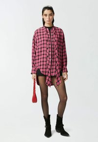 Camicia oversize a scacchi rosa e nera, con bottoni e orlo asimmetrico. Indossata sopra un vestito nero, abbinata a calze nere fino alla coscia e stivali.