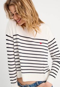 Stribet sweater i creme med marineblå vandrette striber, der har en lille rød hjerte-detalje. Materialet ser blødt ud; designet er afslappet og med løs pasform.
