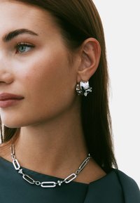 Silverfärgade ringörhängen med dekorativa detaljer, kombinerade med ett lager-på-lager-silverhalsband med förlängda länkar och cirkulära accenter.