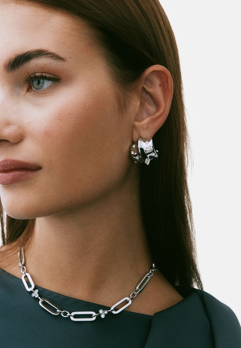 Silverfärgade ringörhängen med dekorativa detaljer, kombinerade med ett lager-på-lager-silverhalsband med förlängda länkar och cirkulära accenter.