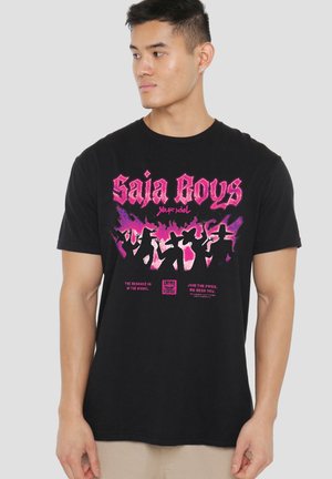 Man wearing black T-shirt with pink graphic text "Saja Boys" and silhouetted figures in hat poses, standing against plain light background.
