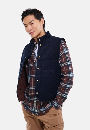 Jeune homme portant un gilet bleu marine boutonné par-dessus une chemise à carreaux rouge et bleue avec un pantalon beige, debout les mains dans les poches, fond clair.