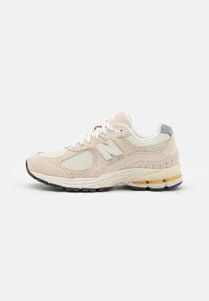 Chaussure de sport beige et blanche avec panneaux en mesh et en suède, fermeture à lacets, semelle rembourrée et semelle extérieure avec bande de roulement noire.