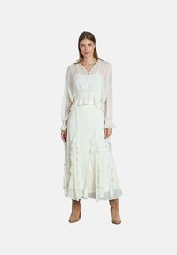 Lungo vestito bianco con volants a strati, colletto petto e vita regolabile con corda. Blusa trasparente a maniche lunghe indossata sopra, abbinata a stivali alla caviglia beige.
