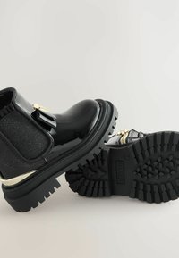 Botas negras de tobillo elegantes con un acabado brillante, lazo decorativo y suelas resistentes, colocadas para mostrar ambos lados y detalles intrincados.