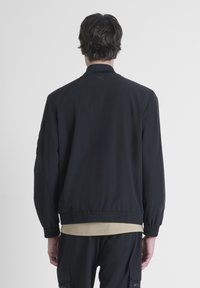 Chaqueta negra con una textura suave, cinturilla elástica y puños acanalados. Presenta un discreto detalle de costura y un pequeño logotipo en la parte posterior.