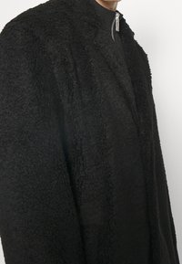 Sort fleecejakke med en tekstureret, shaggy overflade. Har en opretstående krave og en sølvfarvet lynlås. Blødt materiale, oversized pasform.
