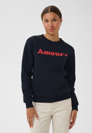 Kvinde med fletninger bærer en sort sweatshirt med rød "Amour" tekst og et hjerte, kombineret med lyse beige bukser, stående på en hvid baggrund.