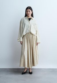 Donna in piedi contro un muro semplice che indossa una camicetta color crema a pois e una gonna lunga coordinata con tacchi neri e orecchini a cerchio dorati.