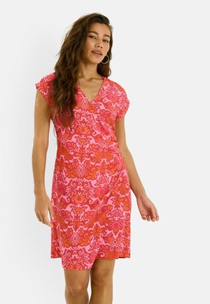 Mujer con pelo largo que lleva un vestido de manga corta con escote en V y un estampado floral rojo y rosa, de pie frente a un fondo blanco.