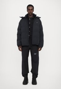 Black puffer jacket with a hood, featuring a quilted texture, worn over a dark checkered shirt and black pants with logo detail. Black boots.