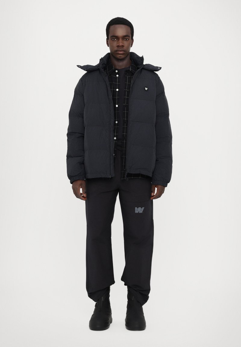 Black puffer jacket with a hood, featuring a quilted texture, worn over a dark checkered shirt and black pants with logo detail. Black boots.