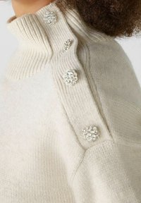 Sweter w kolorze kremowym z prążkowanym stójką, ozdobiony dekoracyjnymi srebrnymi guzikami w kształcie kwiatów wzdłuż szwu na ramieniu. Miękka faktura.