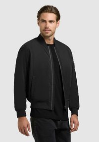 Mann trägt eine schwarze Bomberjacke über einem schwarzen Hemd, steht vor einem hellgrauen Hintergrund und schaut leicht zur Seite.