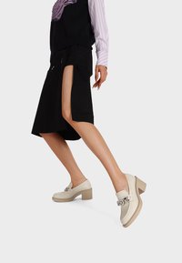 Beige haksloffen met een glanzende afwerking, voorzien van een zilveren kettingaccent en een robuuste hak, tegen een zwarte rok met een zijsplit.