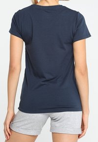 Mujer vistiendo una camiseta de manga corta azul marino lisa y pantalones cortos claros de color gris enrollados, de pie con su espalda hacia el espectador.