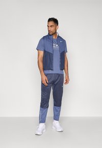 Μπουφάν αμάνικο ναυτικού τύπου πάνω από μπλε μπλούζα με γραφικά, συνδυασμένο με μπλε αθλητικό παντελόνι με υφασμένους τόνους και λευκά αθλητικά παπούτσια.