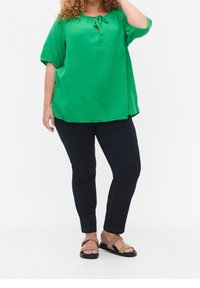 Blusa verde con texture e dettaglio a fiocco sul collo, abbinata a pantaloni in denim scuro e sandali marroni. L'outfit presenta una vestibilità comoda.
