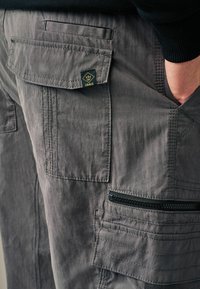 Graue Cargo-Hose mit mehreren Taschen, strukturiertem Stoff und einem schwarzen Reißverschlussdetails. Enthält ein kleines Logo-Patch in Schwarz und Gold.