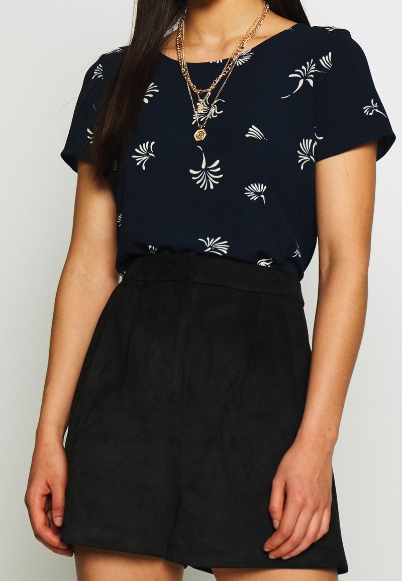 Blouse florale bleu marine à manches courtes, associée à un short noir en suède taille haute. Caractérisée par des motifs blancs délicats et une silhouette élégante.
