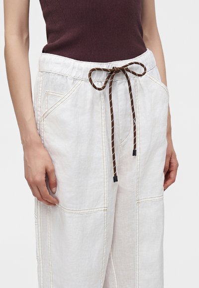Gros plan d'une personne portant un pantalon en lin blanc avec un cordon marron et de grandes poches, associé à un top sans manches marron foncé côtelé.