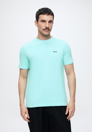 Homme aux cheveux courts et foncés avec une moustache, portant un t-shirt BOSS vert menthe et un pantalon noir sur fond blanc.