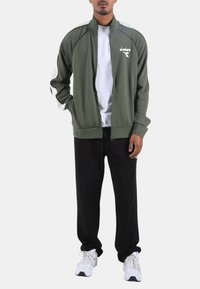 Giacca verde oliva con cerniera, dettagli neri, logo bianco e collo alto abbinata a pantaloni della tuta neri e scarpe da ginnastica bianche.