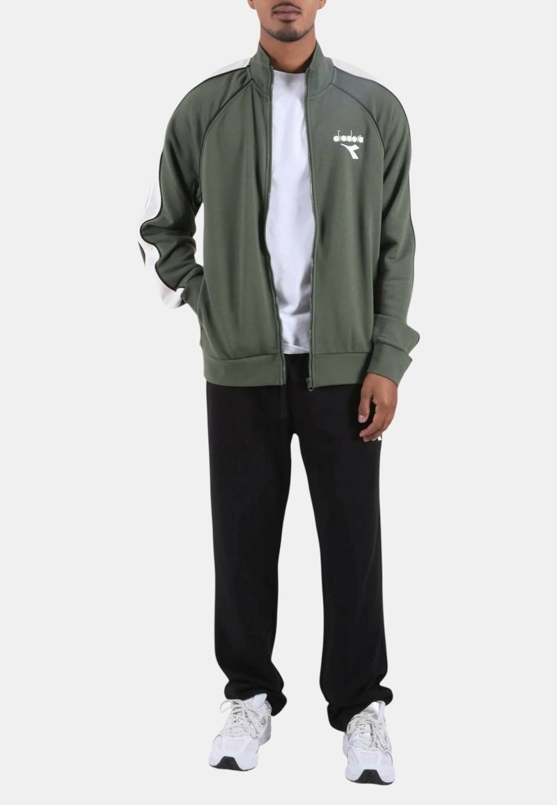 Giacca verde oliva con cerniera, dettagli neri, logo bianco e collo alto abbinata a pantaloni della tuta neri e scarpe da ginnastica bianche.