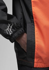 Giacca a vento nera e arancione con polsini elasticizzati, tasche laterali e una distintiva firma bianca sul maniche sinistro.