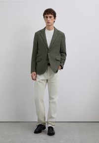 Blazer de patrón espiguilla en verde oliva, combinado con una camisa blanca y pantalones beige claros. Zapatos negros, de pie sobre una superficie gris.