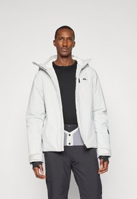 Casaco insulated branco com gola alta, fechamento zip, punhos ajustáveis e um logótipo. Usado sobre uma camisa de manga longa preta e calças cinzentas.
