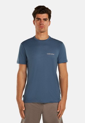 Mężczyzna o krótkich ciemnych włosach, ubranony w niebieski t-shirt Icebreaker i beżowe spodnie, stojący na jednolitym jasnym tle.