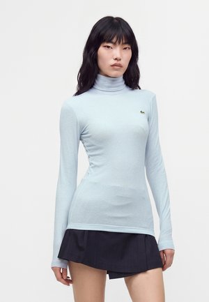 Mujer con cabello largo y negro lleva un suéter de cuello alto azul claro y una falda corta negra, de pie frente a un fondo blanco liso.