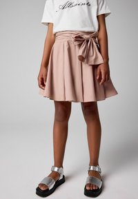 Falda plisada de color rosa pálido con detalle de cintura atada, combinada con sandalias planas plateadas, con una plataforma negra y una sola correa con hebilla.